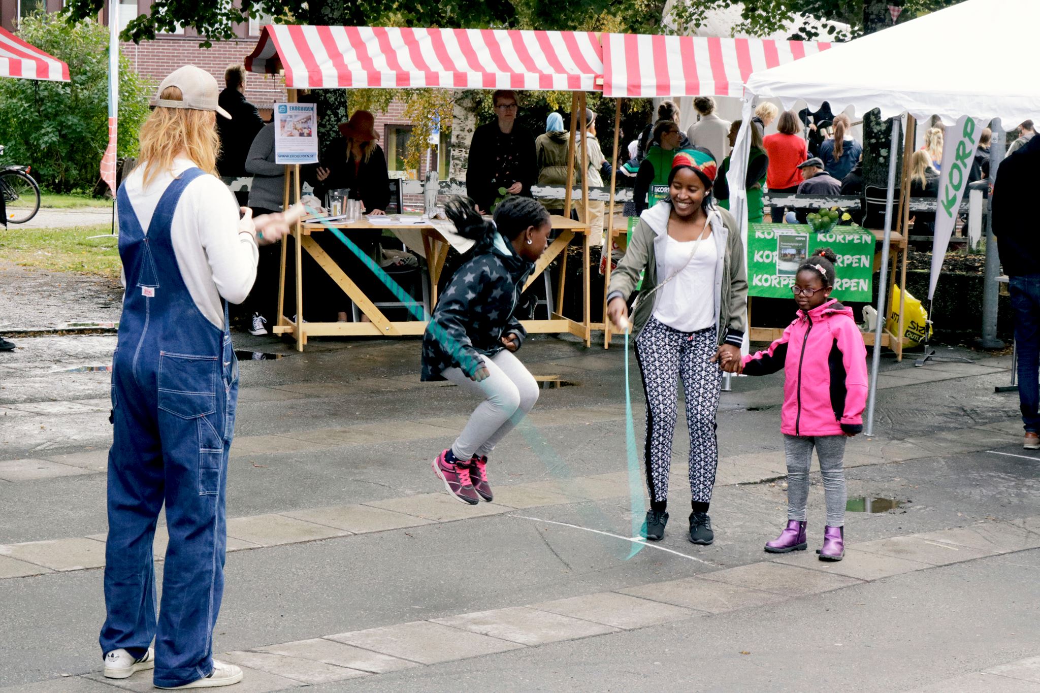 En bild på en liten flicka som hoppar hopprep, två vuxna håller repet. Bakom skymtas en marknadsplats