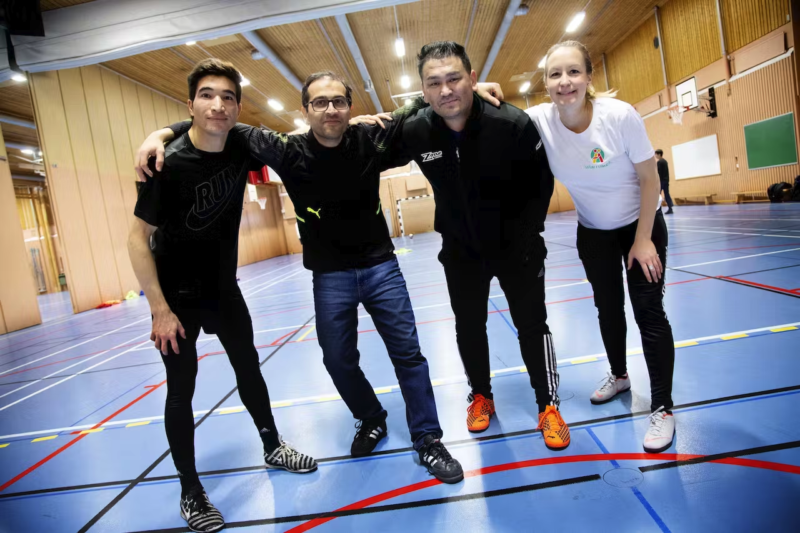 Fyra personer står på en inomhusfotbollsplan . De håller armarna om varandra och ser glada ut. En kvinna, fyra män.